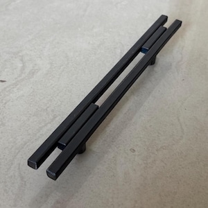 May include: Black metal cabinet pulls with a modern, minimalist design. The pulls are rectangular and have a matte finish. The hardware is suitable for drawers or cabinet doors.