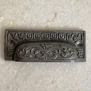 May include: A black cast iron drawer pull with an ornate design. The pull has a rectangular shape with a curved center section. The design features a repeating pattern of squares and a floral motif.