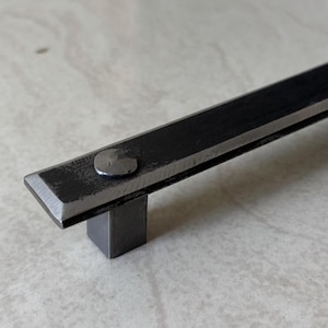 Op de afbeelding: Close-up van een donkergrijze metalen kastgreep. De rechthoekige handgreep heeft een vierkante basis en is bevestigd met een enkele klinknagel. Het metaal heeft een getextureerd, industrieel uiterlijk. De greep is ontworpen voor een kast of lade.