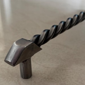 以下が含まれることがあります： 手作りの、ねじれたバーのデザインの暗い金属製のキャビネットプル。ハンドルは幾何学的な端部と、取り付け用の2つの円筒形のポストを備えています。全長は約25cmです。