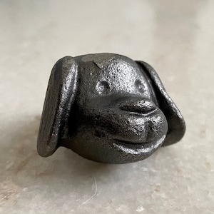 Op de afbeelding: Een zwarte gietijzeren deurknop in de vorm van een hondenkop met een glimlachende uitdrukking.