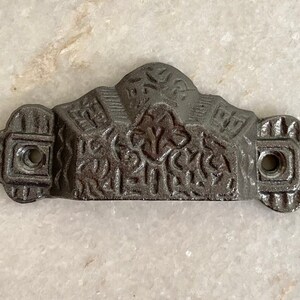 May include: A grey metal decorative hardware piece with a floral design and two screw holes. The piece has a curved top and a flat bottom.
