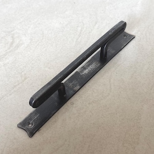 May include: A black metal door pull with a rectangular backplate and a rounded handle. The handle is attached to the backplate with two curved supports. The door pull has a distressed finish, giving it a rustic appearance.