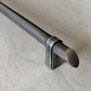 Puede incluir: Primer plano de un tirador de armario metálico con acabado gris oscuro. El tirador es cilíndrico con extremos redondeados y está fijado a soportes rectangulares. El herraje tiene una estética rústica e industrial.