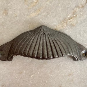 May include: A cast iron shell-shaped drawer pull with a textured surface. The hardware is a dark grey colour and has two holes for mounting. This decorative pull is suitable for cabinets and drawers.