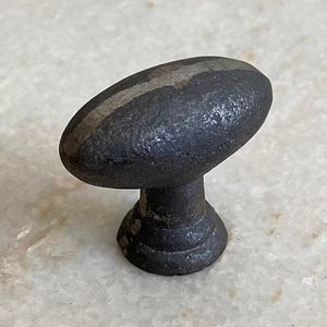 May include: A dark grey oval cabinet knob with a lighter grey stripe down the centre. The knob has a textured surface and a short, rounded base. The hardware is likely made of metal and is designed for furniture.