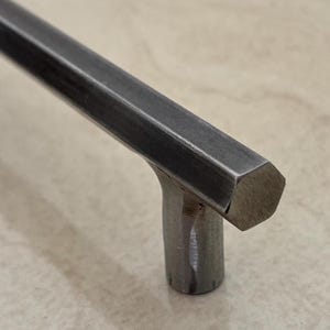 Puede incluir: Primer plano de un tirador de armario de metal cepillado con un mango hexagonal. El mango está conectado a una base cilíndrica. El herraje tiene un acabado mate y está colocado sobre un fondo neutro.