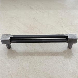 Puede incluir: Un tirador de metal para armario con una barra rectangular y dos ranuras paralelas. Los extremos son cuadrados con un poste cilíndrico que se extiende hacia abajo. El acabado es de color plata mate.