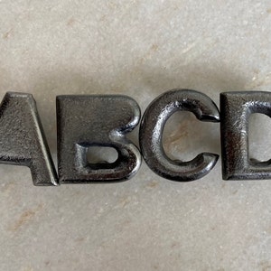 Puede incluir: Cuatro letras de metal, A, B, C y D, están dispuestas en fila sobre una superficie gris claro. Las letras son de metal gris oscuro y tienen una textura ligeramente rugosa.