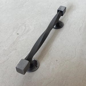 Peut inclure: Une poignée de porte en fer forgé gris foncé avec un design de barre torsadée. La poignée a des embouts carrés et des plaques de montage circulaires. La quincaillerie est conçue pour une esthétique rustique ou industrielle.