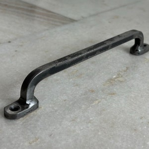 May include: A black metal drawer pull with a simple, curved design. The pull has a slightly distressed finish, giving it a rustic look.
