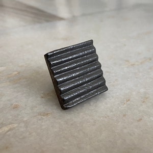 Może przedstawiać: Mały, kwadratowy, czarny metalowy przedmiot z fakturowaną, falistą powierzchnią.