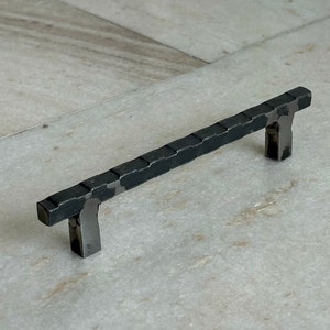 Puede incluir: Tirador de armario de metal negro con acabado martillado. El tirador mide aproximadamente 10 cm de largo.