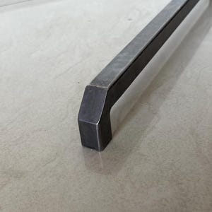 May include: A close-up of a dark grey metal cabinet pull with a rectangular bar and a squared-off handle. The handle is angled and has a solid, industrial design. The pull is likely for a drawer or cabinet.