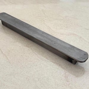 Könnte beinhalten: Ein rechteckiger, dunkelgrauer Metall-Schubladengriff mit abgerundeten Enden. Der Griff hat eine lange, schlanke Form und ist für die horizontale Montage konzipiert. Das Metall hat ein leicht strukturiertes Aussehen, das auf eine gebürstete oder gealterte Oberfläche hindeutet.