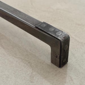 Może przedstawiać: Zbliżenie metalowego uchwytu z kątem prostym. Uchwyt wykonany jest z ciemnego, prawdopodobnie zardzewiałego metalu, z widocznymi śladami spawania. Projekt jest przemysłowy i minimalistyczny.