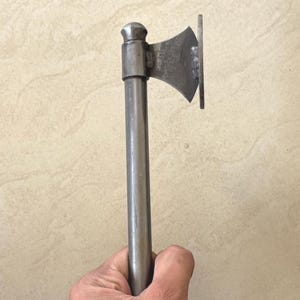 May include: A small metal axe with a rounded top and a flat, rectangular base. The axe head is curved with a sharp edge. The cylindrical handle is made of the same metal as the head. The axe is held in a hand, with a light-colored background.