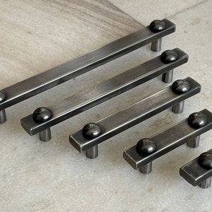 May include: Five different sizes of black metal drawer pulls with rounded ends. The pulls are arranged in a staggered pattern from largest to smallest.