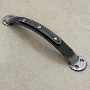 Könnte beinhalten: Ein verwitterter, dunkelgrauer Metallgriff mit geschwungenem Design. Der Griff hat an jedem Ende zwei kreisförmige Montageplatten mit drei Schraubenlöchern. Der Mittelteil hat ein genietetes Aussehen.