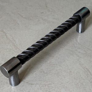 May include: A metal cabinet pull with a twisted, textured center and cylindrical end pieces. The hardware is a dark gray color, and the design is industrial.