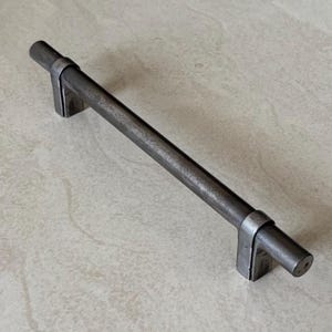 May include: A dark grey metal cabinet pull with a cylindrical bar and rectangular mounting brackets. The handle is approximately 20 cm long, with a textured, industrial finish. Suitable for furniture restoration or modern design.