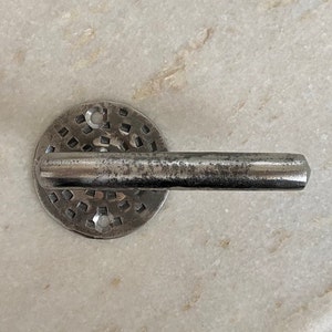 Peut inclure: Une poignée de porte en métal argenté avec une base ronde décorative présentant un motif en grille. La poignée est légèrement incurvée et présente une finition usée et vieillie.