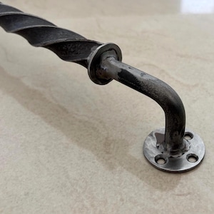 Peut inclure: Une poignée de porte en fer forgé avec un design torsadé et texturé. La poignée est gris foncé avec un bras incurvé et une plaque de montage circulaire avec quatre trous. Le métal a une finition vieillie.