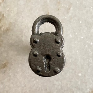 Può includere: Un piccolo lucchetto in stile antico con finitura metallica scura. Il lucchetto presenta una maniglia superiore arrotondata e una serratura, con rivetti decorativi attorno al corpo. Il design suggerisce un'estetica vintage o rustica.