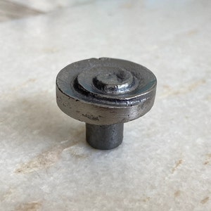 Op de afbeelding: Een donkergrijze metalen kastknop met een spiraalvormig ontwerp op de ronde kop. De knop heeft een korte, cilindrische steel. De afwerking lijkt enigszins verweerd, wat hem een rustieke uitstraling geeft. De knop is ontworpen voor meubels.