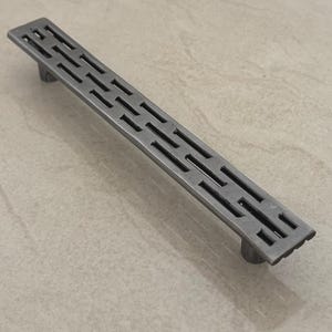 Op de afbeelding: Een rechthoekige kastgreep in antieke stijl met een geometrisch ontwerp. De metalen greep heeft een gedempte grijze kleur en heeft een reeks rechthoekige uitsparingen. Aan elk uiteinde bevinden zich twee afgeronde handgrepen.
