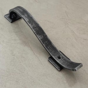 Puede incluir: Un tirador de puerta de metal gris oscuro desgastado con un asa curvada. El asa está unida a dos placas de montaje cuadradas. El metal tiene una apariencia texturizada y envejecida, lo que sugiere un estilo vintage o rústico. El tirador mide unos 20 cm de largo.