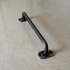 Op de afbeelding: Een industriële deurklink van donkergrijs metaal. De gebogen handgreep heeft een ruwe textuur en bevestigingsplaten met schroefgaten. Het gesmede uiterlijk suggereert een handgemaakt ontwerp. De lengte is ongeveer 15 cm.