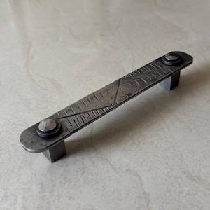 Könnte beinhalten: Handgefertigter Schrankgriff aus dunkelgrauem Metall. Der rechteckige Griff hat eine strukturierte Oberfläche mit geätzten Linien und ist mit quadratischen Pfosten mit nietartigen Details verbunden. (ca. 15 cm)