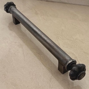May include: A rustic, dark metal door pull with a cylindrical bar and decorative, flower-shaped ends. The handle has two rectangular mounting brackets. The metal has a textured, aged appearance.