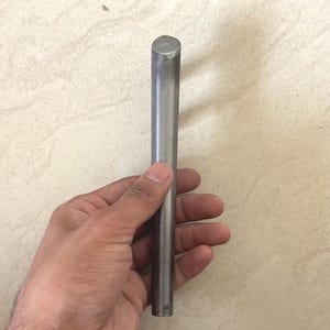 May include: A cylindrical, silver metal rod with a flat top and bottom. The rod is held in a hand against a neutral background. The metal appears unpainted and has a smooth, reflective surface.