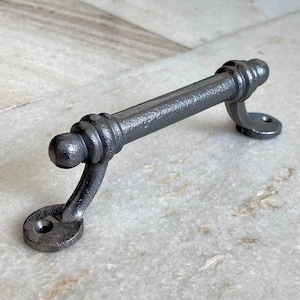 Może przedstawiać: Srebrny, żeliwny uchwyt do drzwi o zaokrąglonym kształcie. Uchwyt ma gładkie, fakturowane wykończenie i jest przeznaczony do użytku na drzwiach i szafkach.