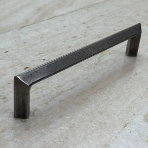 Puede incluir: Un tirador de armario rectangular gris oscuro con un perfil cuadrado. El tirador está hecho de metal y tiene una superficie ligeramente texturizada. El tirador está diseñado con ángulos rectos y es adecuado para cajones o armarios.
