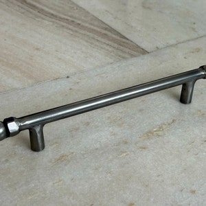 以下が含まれることがあります： 円筒形のバーとボール型の端が付いた、つや消し金属製のキャビネットプル。ハンドルには2つの垂直サポートがあります。ハードウェアは濃いグレー色で、家具用に設計されています。プルは約15cmの長さです。