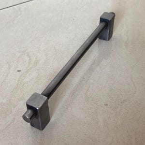 May include: A dark gray metal cabinet pull with a rectangular handle and square end pieces. The handle is a straight rod connecting the square ends. The hardware is designed for use on drawers or cabinet doors.