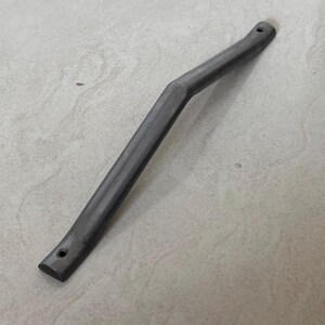 Puede incluir: Un asa de metal curvada de color gris oscuro con dos agujeros en los extremos. El asa tiene una ligera curva en el centro y parece estar hecha de un material sólido y duradero. El objeto es probablemente un componente de un artículo más grande.