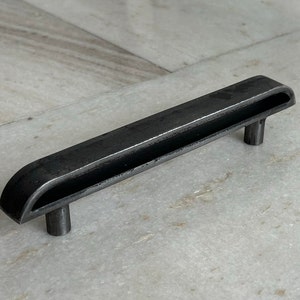 Könnte beinhalten: Schwarzer Metall-Schubladengriff mit rechteckiger Form und einem Ausschnitt in der Mitte. Der Griff hat abgerundete Kanten und eine leicht strukturierte Oberfläche.