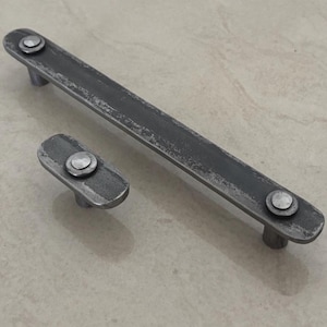 Könnte beinhalten: Ein Set aus zwei verwitterten, dunkelgrauen Metall-Schrankgriffen. Einer ist ein langer, rechteckiger Balken mit zwei Griffen, der andere ein kleinerer, ovaler Griff mit einem Griff. Beide haben ein Used-Finish und sichtbare Nieten.