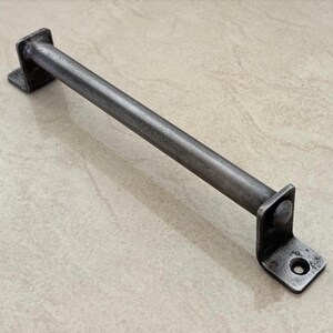Könnte beinhalten: Ein Metall-Türgriff mit einem rechteckigen Stab und quadratischen Montageplatten. Der Türgriff besteht aus dunkelgrauem Metall mit leicht strukturierter Oberfläche. Die Platten haben ein Loch für Schrauben.