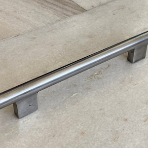 Op de afbeelding: Een zilveren metalen kastdeurgreep met een rechthoekige basis en een afgeronde stang.