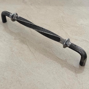 Peut inclure: Une poignée de porte en fer forgé avec une partie centrale torsadée et des extrémités incurvées. La poignée est gris foncé avec des accents argentés. La quincaillerie est conçue pour une porte ou un meuble.