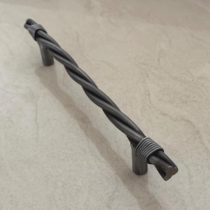 Puede incluir: Un tirador de armario de metal gris oscuro retorcido con un diseño similar a una cuerda. El tirador está soportado por dos postes cilíndricos cortos. El diseño presenta una estética industrial texturizada, adecuada para muebles o armarios.