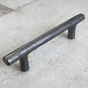 Op de afbeelding: Een zwarte metalen kastgreep met een ruwe, getextureerde afwerking. De greep is ongeveer 10 cm lang en heeft een eenvoudig, modern ontwerp.