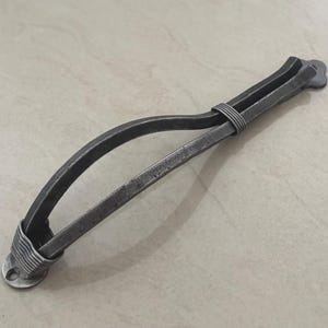 Puede incluir: Tirador de armario de metal gris oscuro forjado a mano, con un asa curvada y un envoltorio decorativo de alambre. El tirador presenta una base plana y circular y una pieza final rectangular. Esta ferretería rústica añade un toque único a los muebles.