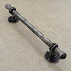 Puede incluir: Un tirador de puerta de metal rústico con un acabado envejecido. El tirador es una barra recta con extremos redondeados, conectada a placas de montaje circulares. La ferretería es de color gris oscuro.