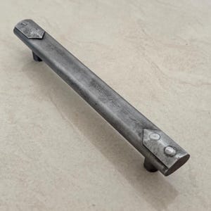 Puede incluir: Un tirador de armario de metal gris envejecido, con una barra rectangular y dos pequeños tiradores redondos. Los extremos presentan placas metálicas decorativas con remaches, añadiendo una estética industrial rústica. El tirador mide unos 15 cm de largo.